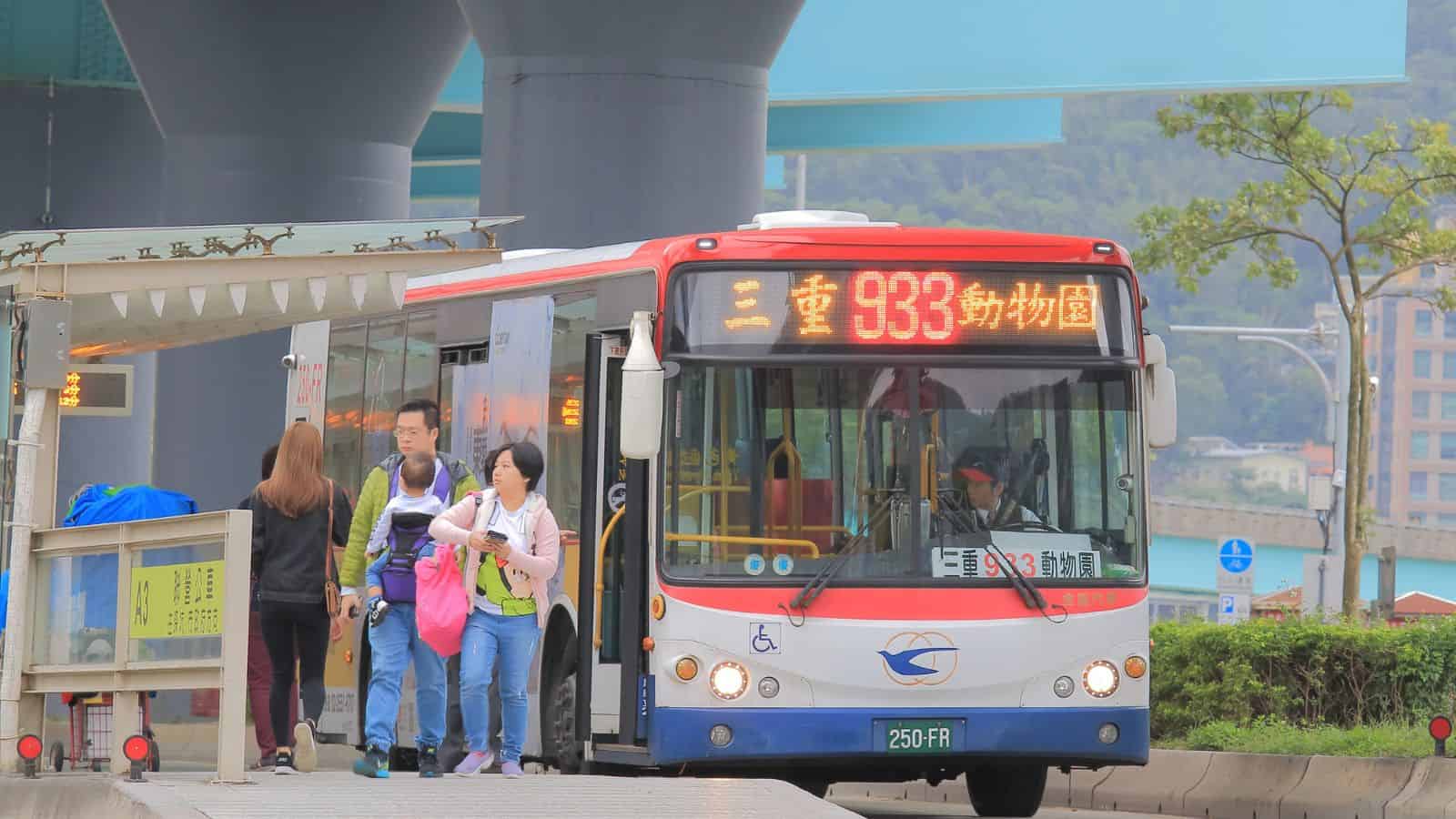 People boarding and exiting a city bus displaying route 933 at a bus stop under a highway overpass.