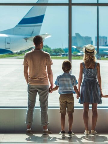 A family of four stands at an airport window, watching airplanes on the runway.