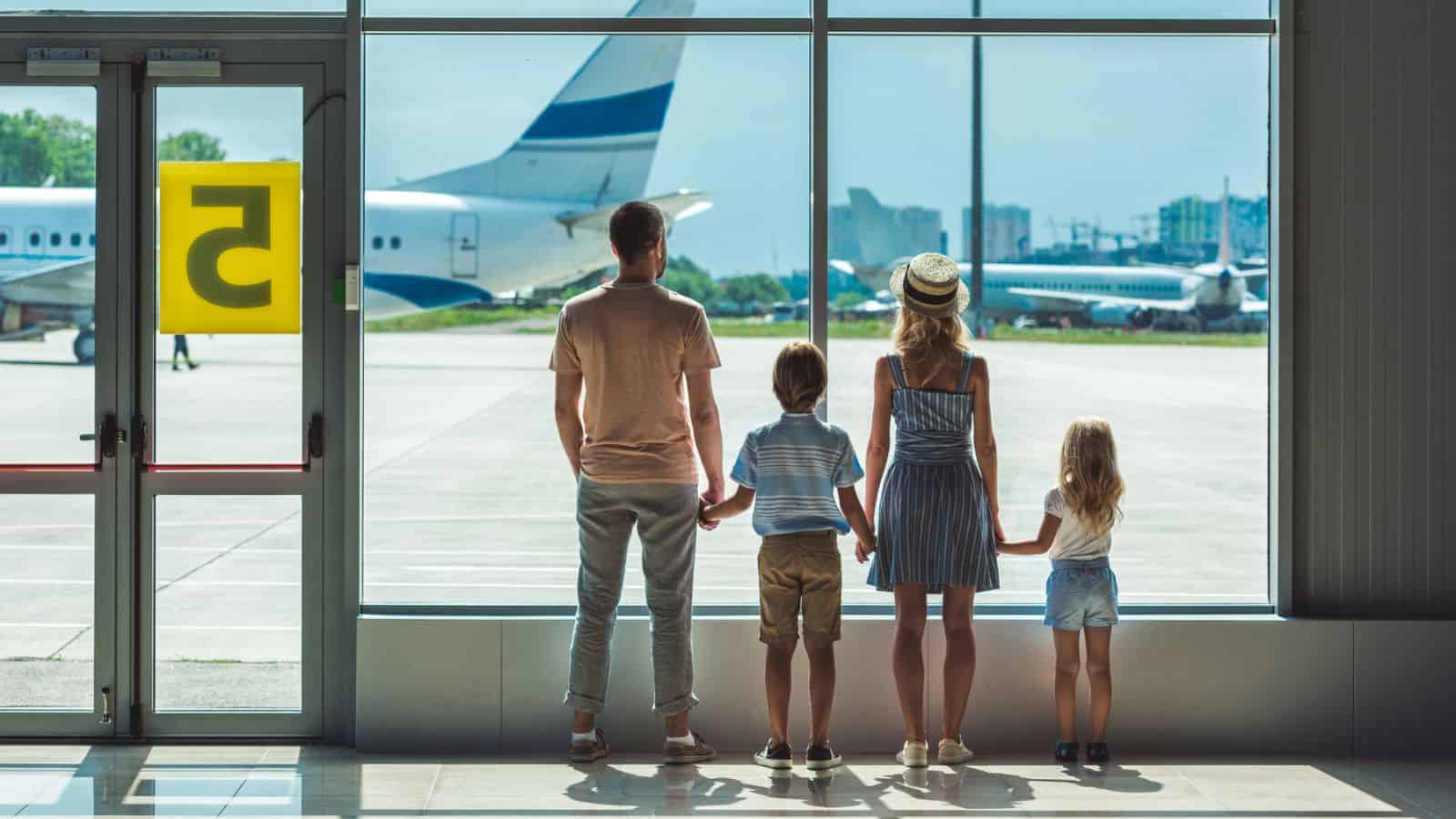 A family of four stands at an airport window, watching airplanes on the runway.