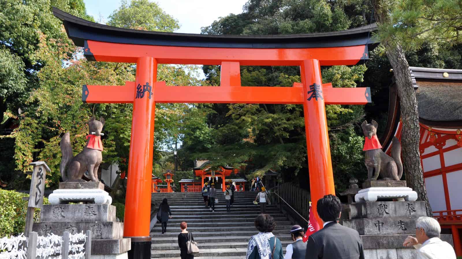 People walk up stone steps under a large red torii gate with fox statues at a Japanese shrine.