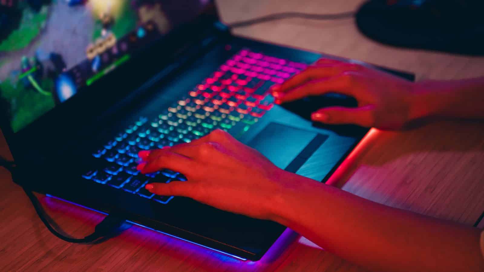 Hands playing a video game on a laptop with a colorful, illuminated keyboard.