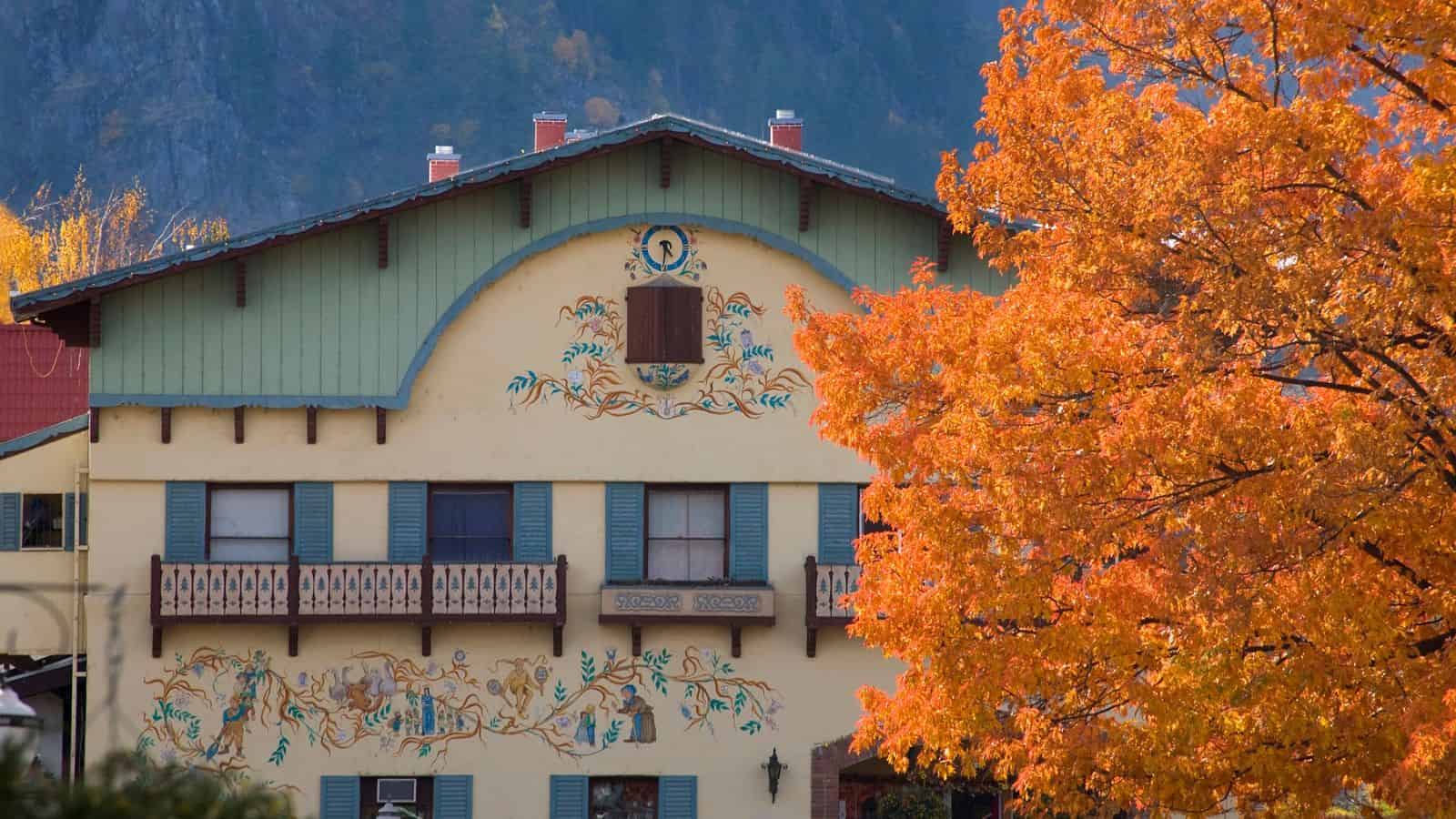 Bavarian-style building with murals and a balcony, next to a bright orange autumn tree.