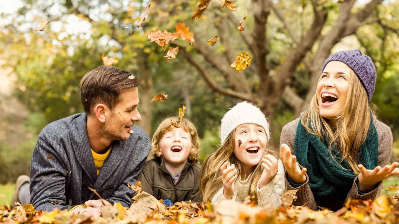 A family of four lying in fallen leaves, smiling and playing together in an autumn park.