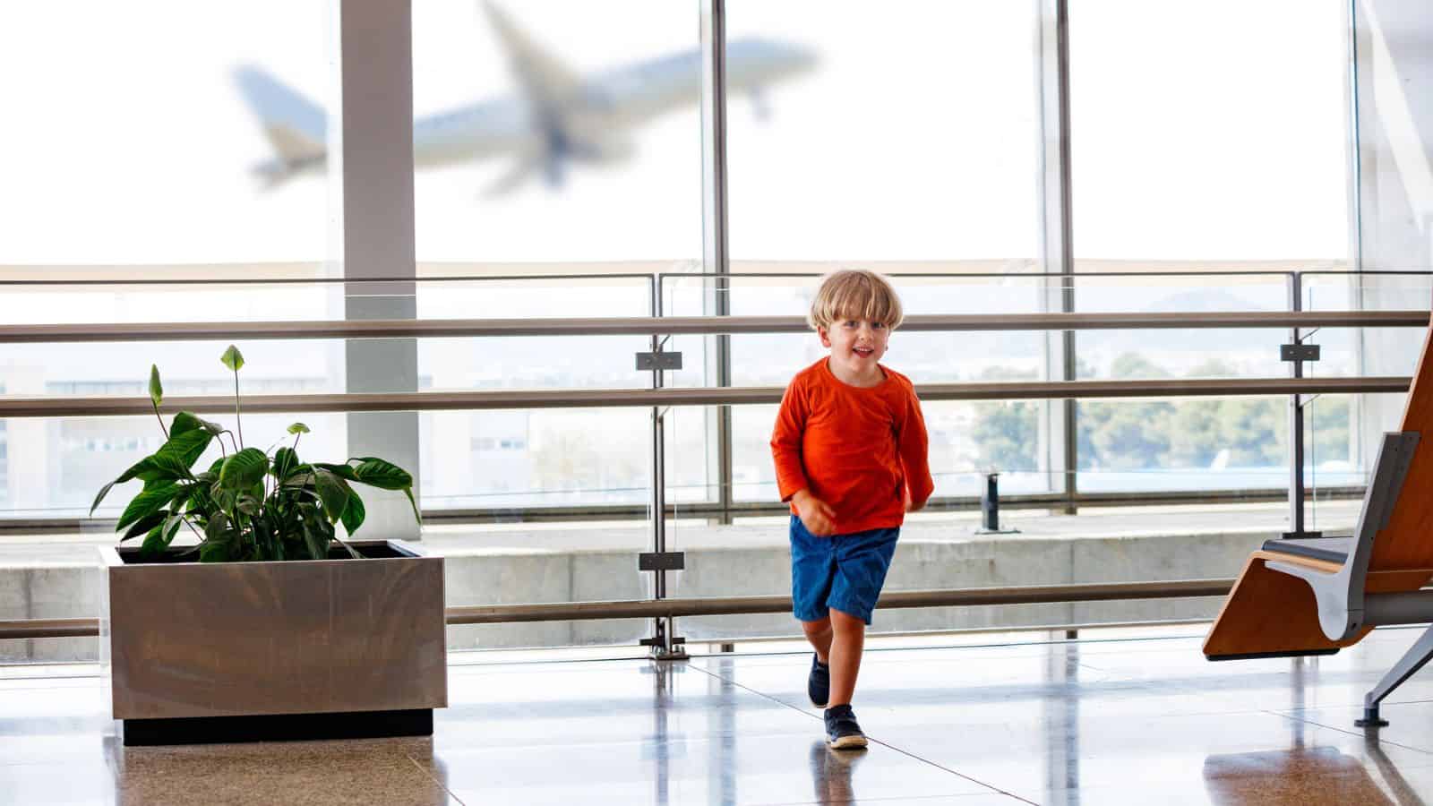 Young boy in orange shirt running in airport terminal with a blurred airplane seen through the window behind him.