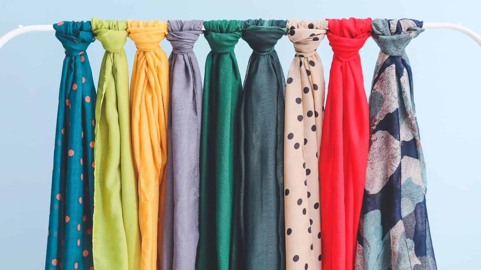 Eight colorful scarves in various solid colors and patterns hang neatly on a white rack against a blue background.