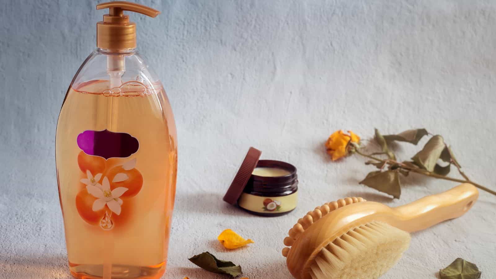 A bottle of liquid soap, a wooden brush, a small cream jar, and dried flowers on a light surface.
