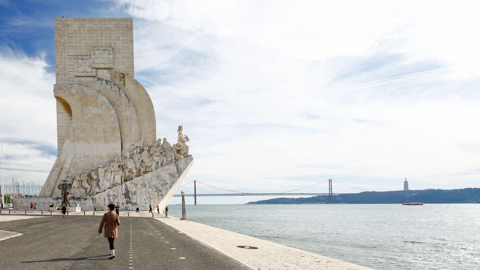 People walking by the Monument to the Discoveries near the Tagus River in Lisbon, Portugal, with a bridge in view.