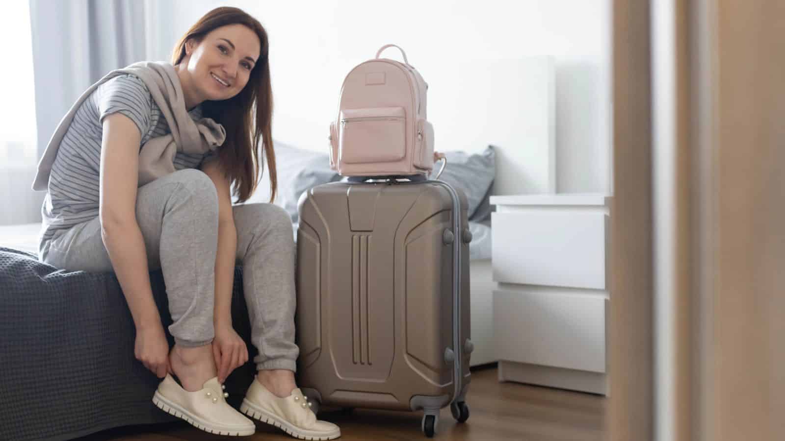 Woman sitting on a bed, putting on shoes next to a suitcase and a backpack in a bedroom.