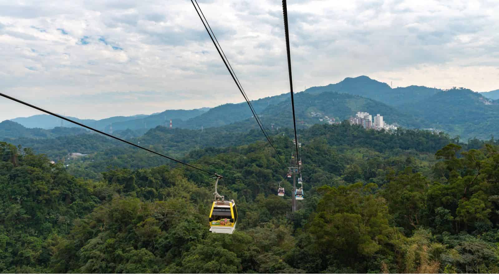 A cable car travels over lush green hills with mountains and buildings in the background under a cloudy sky.