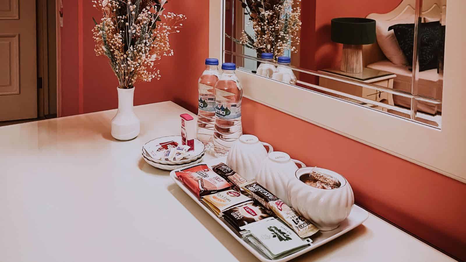 A white table with snacks, tea bags, water bottles, cups, and a vase with flowers beside a mirror.