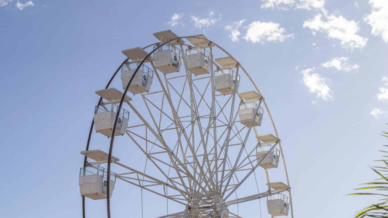 White Ferris wheel with empty cabins against a bright blue sky with a few clouds.