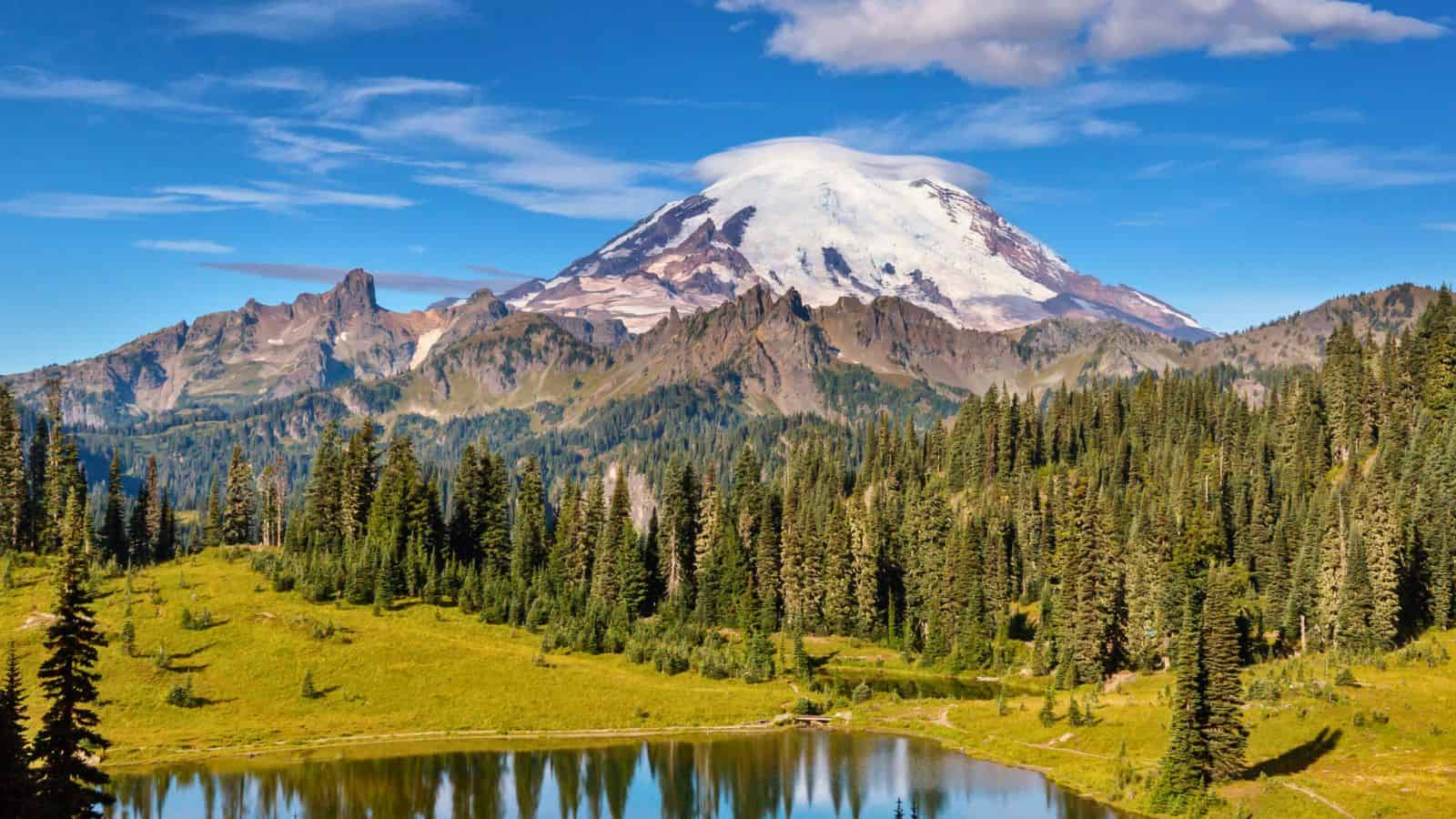 Snow-capped mountain with green forest and a reflective lake under a blue sky with scattered clouds.