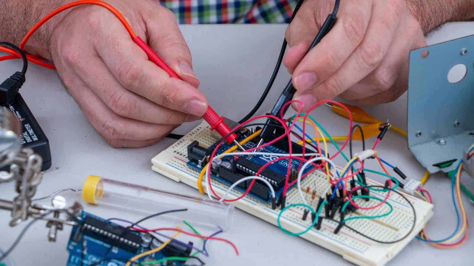 Hands using probes to test an electronic circuit with wires and a breadboard on a table, similar to devices that might raise airport security concerns if stopped at an airport in 2025.