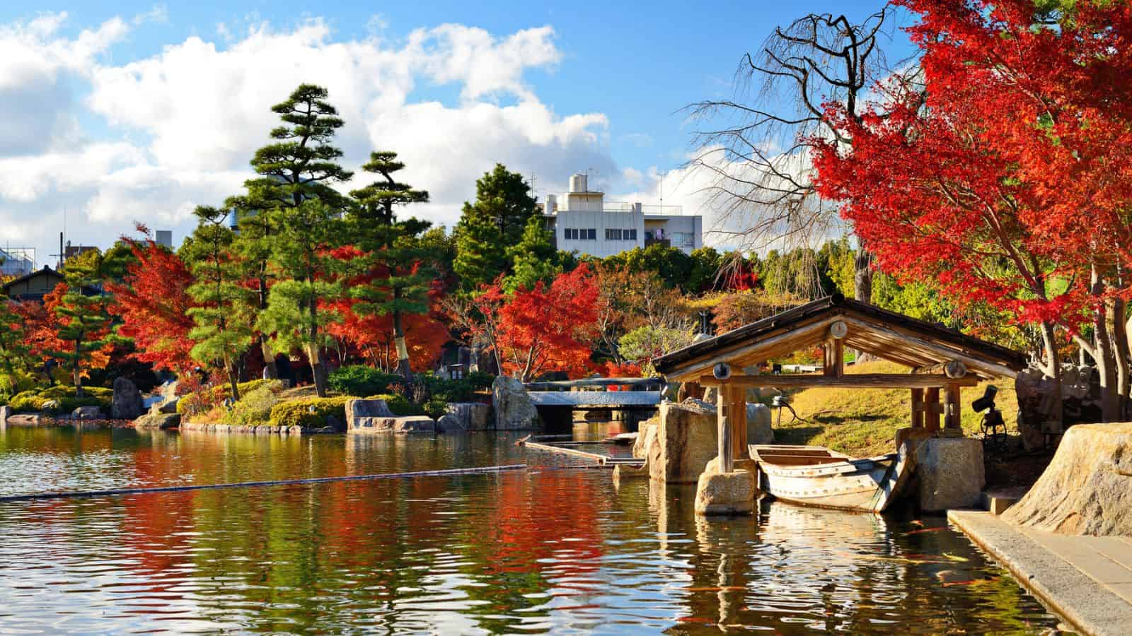 A serene Japanese garden with a pond, red autumn trees, a wooden shelter, and boats under a blue sky.