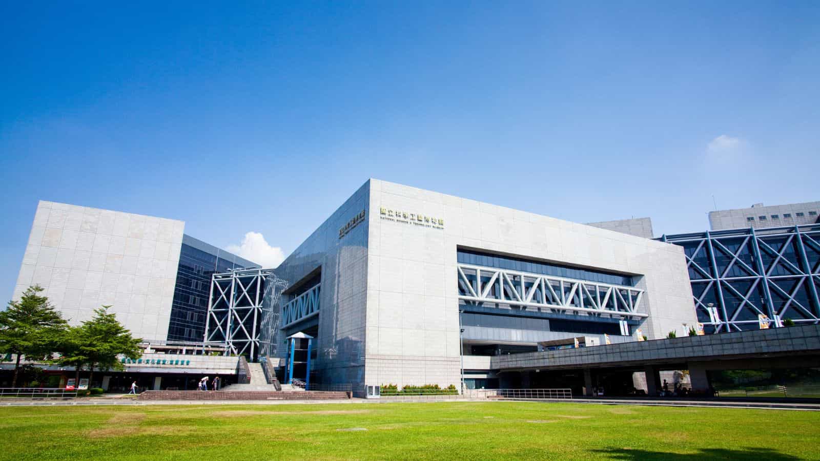 A large modern gray building with geometric design stands under a clear blue sky and is surrounded by green grass.