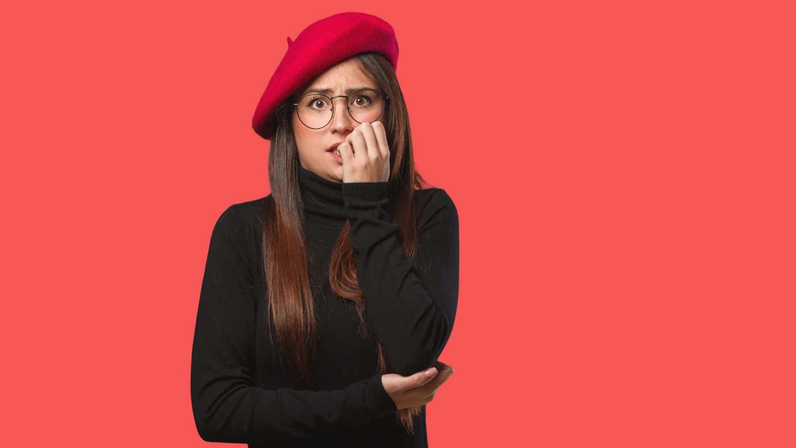 Woman in a red beret and black top looking anxious, biting her nails against a solid red background.