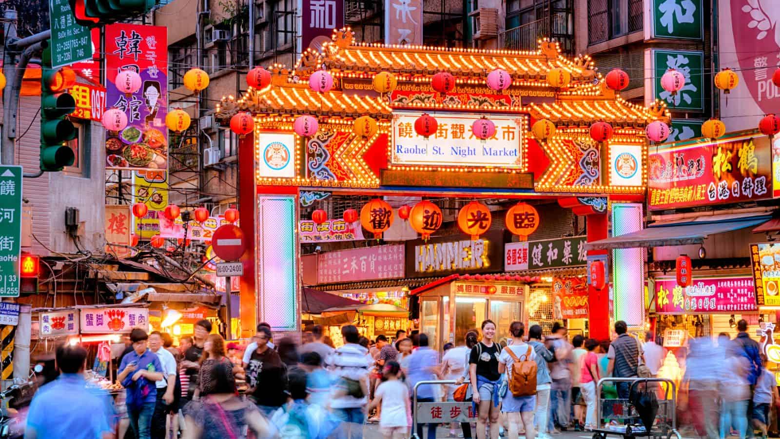 Crowds walk past bright neon lights and lanterns at the entrance to Raohe St. Night Market in Taipei, Taiwan.