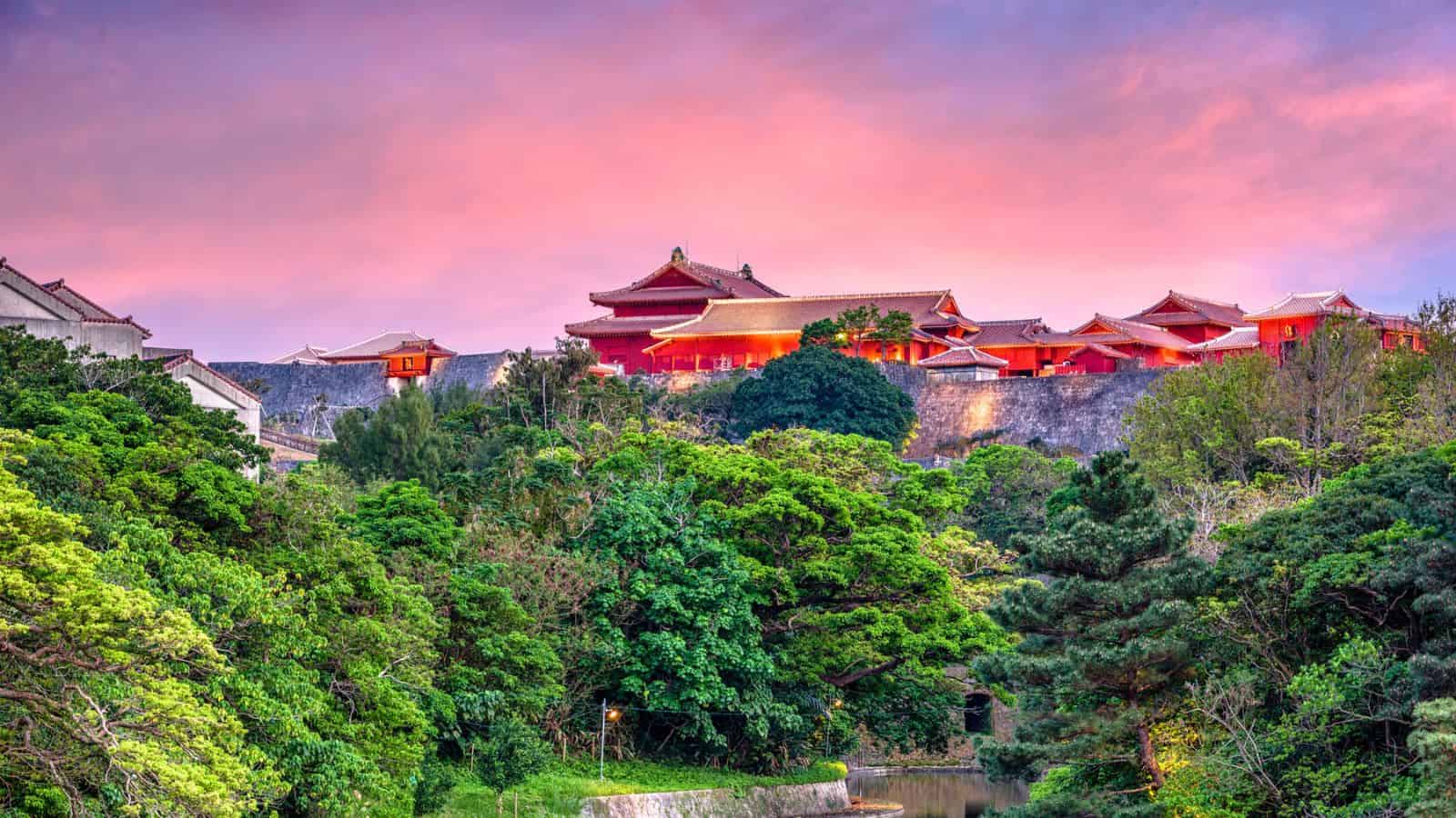 Shuri Castle in Okinawa, Japan, surrounded by lush trees under a vibrant pink and purple sky at sunset.