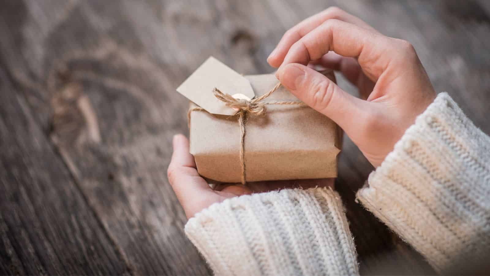 Hands in a white sweater hold a small, gift-wrapped box tied with twine and a blank tag on a wooden surface.
