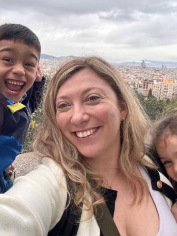 A smiling woman takes a selfie with two happy children, with a cityscape in the background.