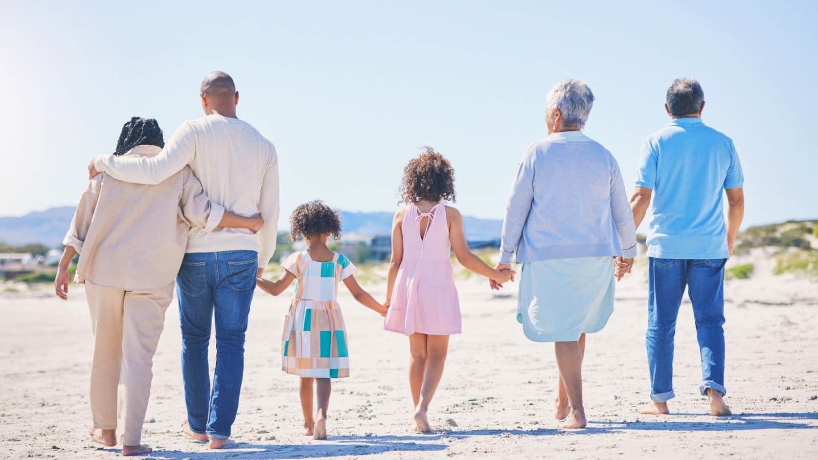 Three adults and three children walk hand-in-hand on a sunny beach, seen from behind.