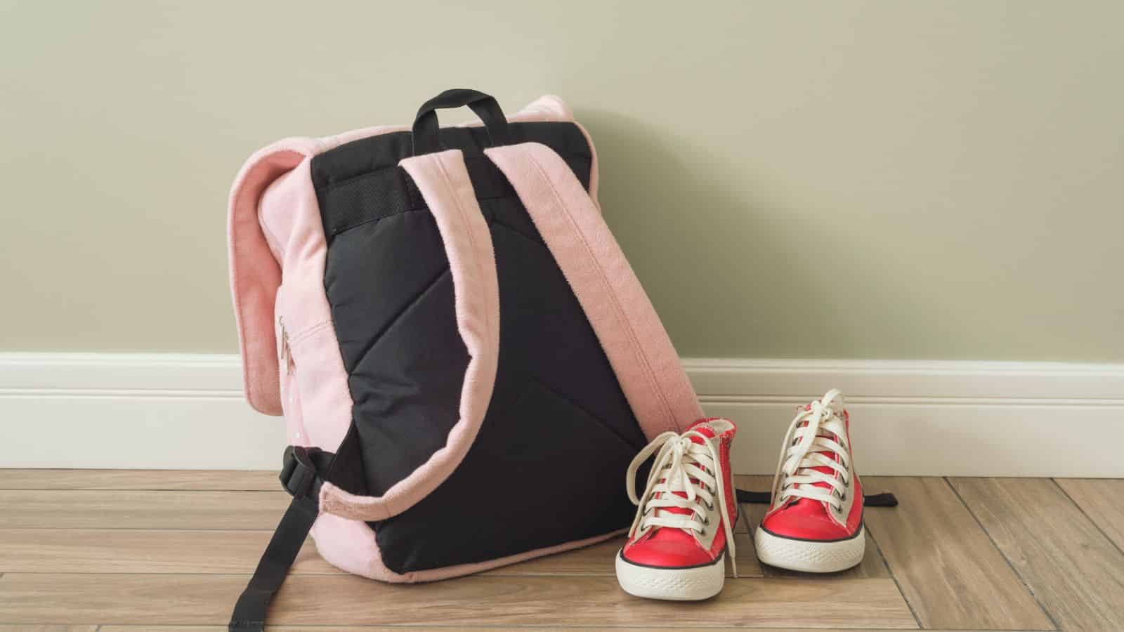 A pink and black backpack next to a pair of red and white sneakers on a wooden floor.