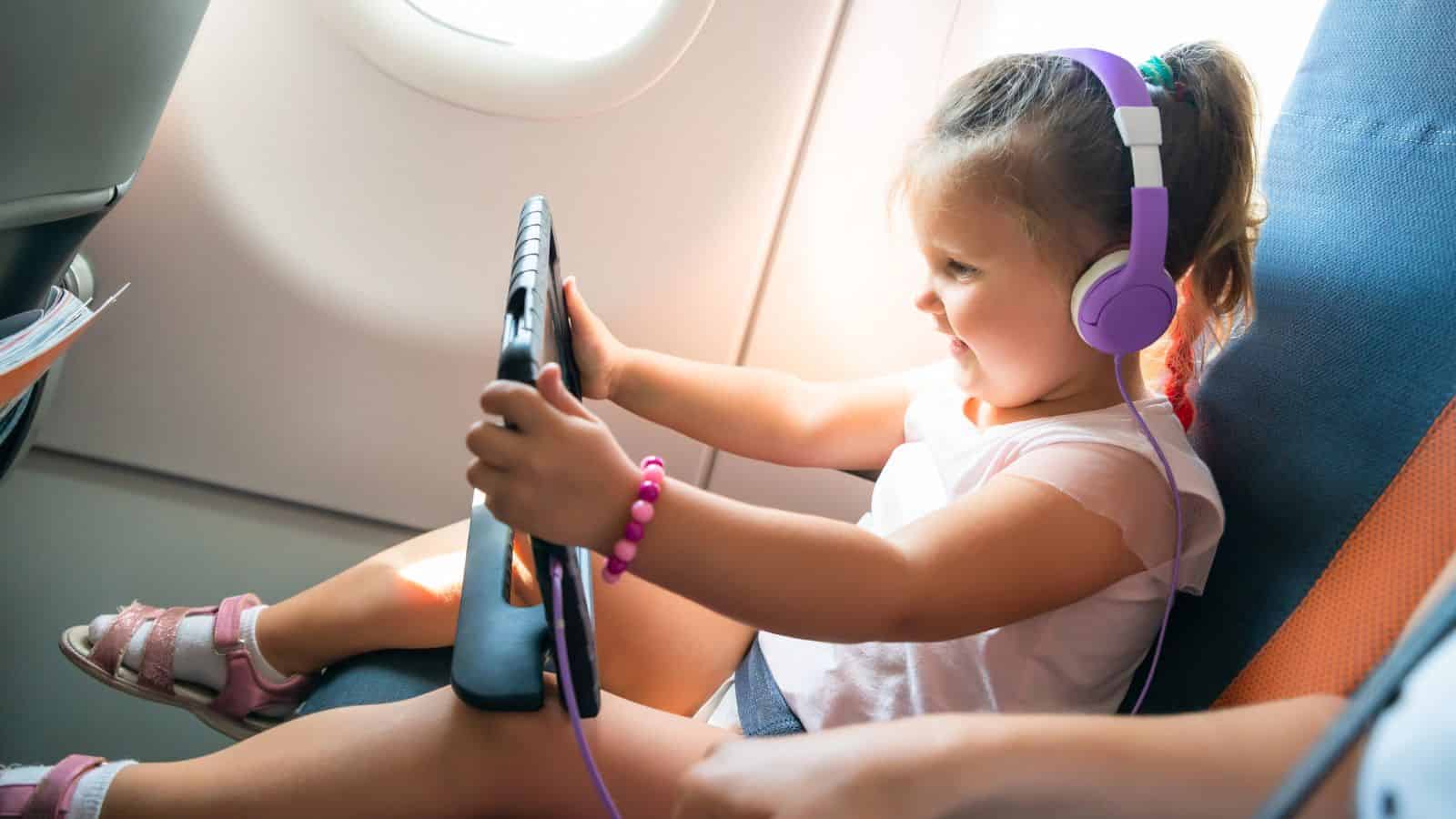 Young girl wearing purple headphones, smiling and using a tablet while sitting in an airplane seat.