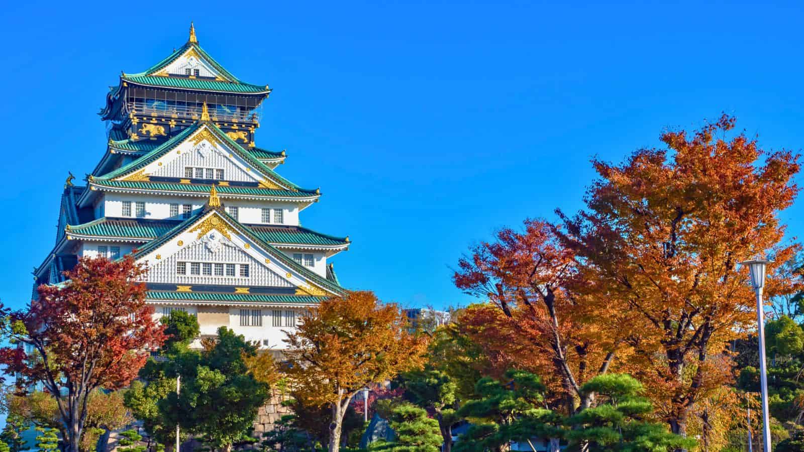 Osaka Castle with green and gold roof, surrounded by colorful autumn trees under a clear blue sky.