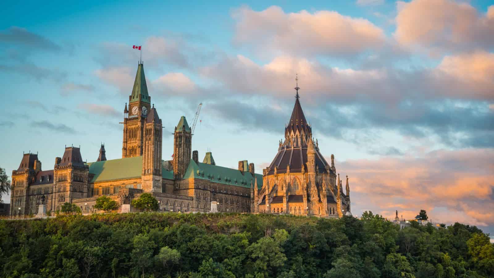 Canadian Parliament buildings in Ottawa at sunset, with green roofs and a Canadian flag, viewed from across trees.