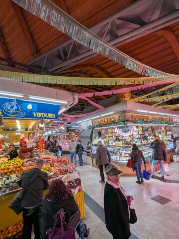 People shopping at an indoor market with colorful fruit and vegetable stalls under a wooden arched ceiling—a perfect spot to discover fresh food and experience the vibrant atmosphere of Barcelona markets.