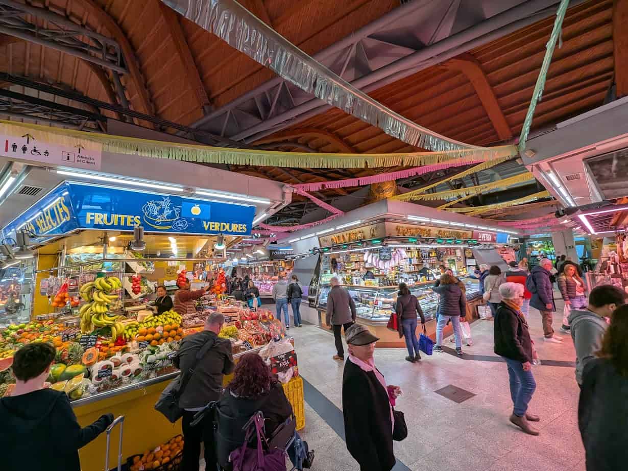 People shopping at an indoor market with colorful fruit and vegetable stalls under a wooden arched ceiling—a perfect spot to discover fresh food and experience the vibrant atmosphere of Barcelona markets.