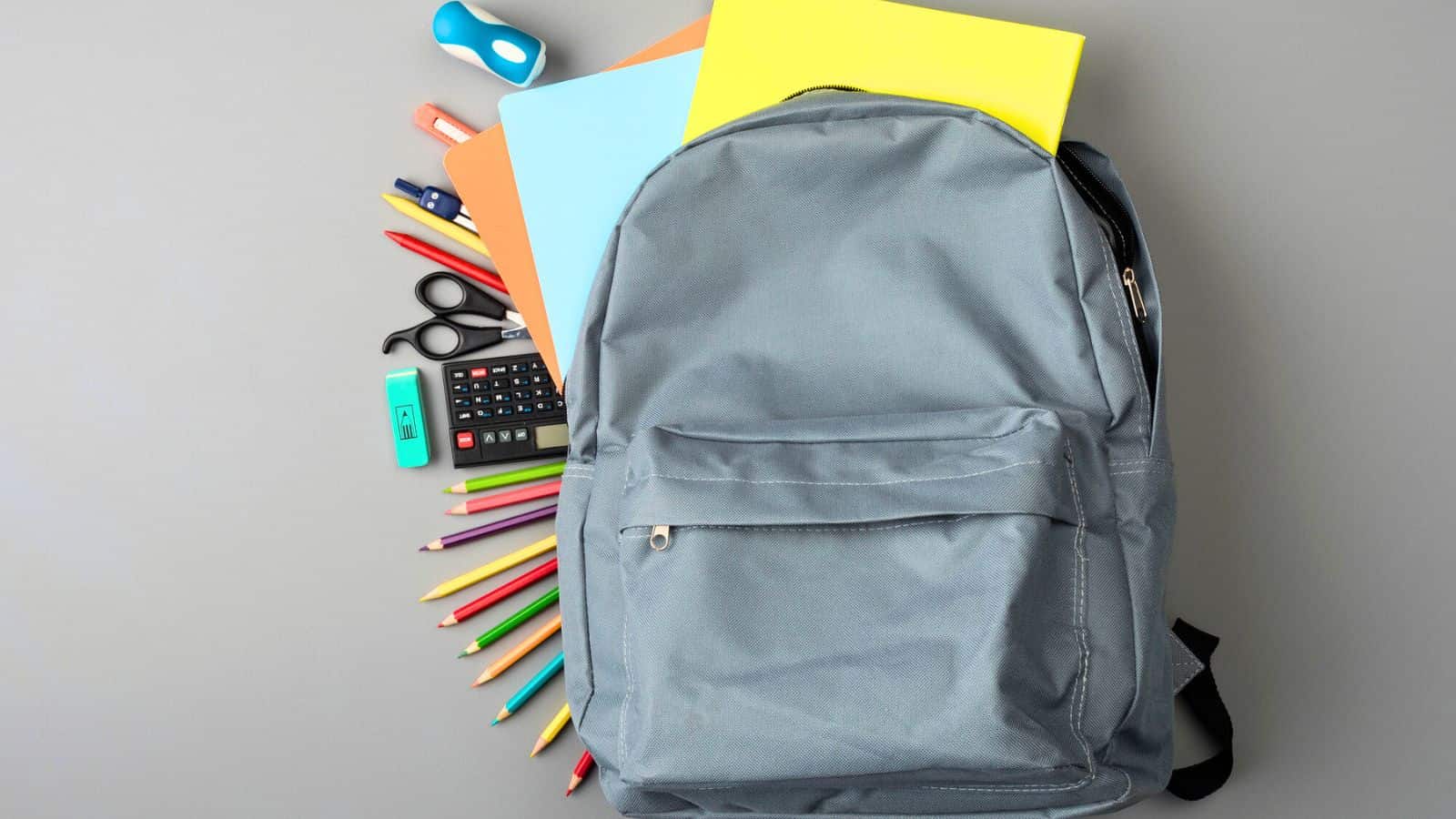 Gray backpack with colored pencils, notebooks, scissors, ruler, calculator, and other school supplies on a gray surface.