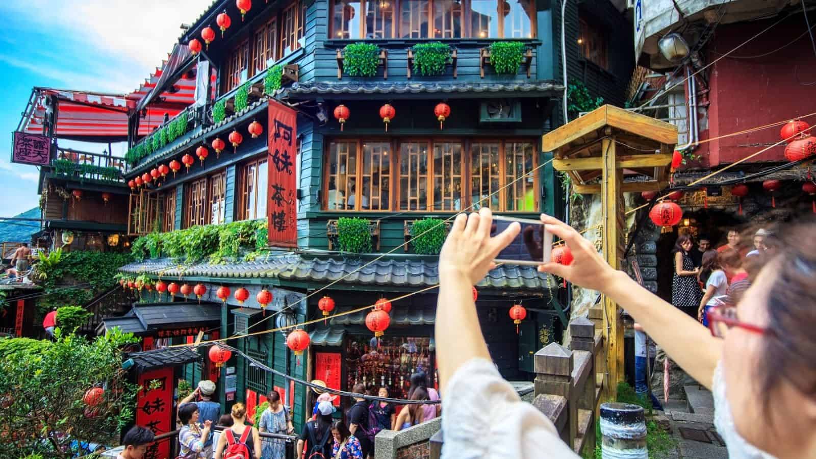 Person taking a photo of a lantern-lit, multi-story teahouse in a bustling, colorful street scene.