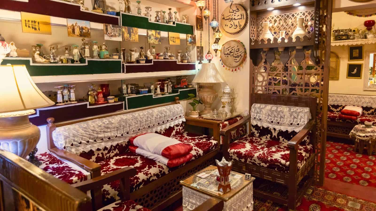 A cozy traditional Middle Eastern living room with ornate decor, patterned sofas, shelves, and decorative items.