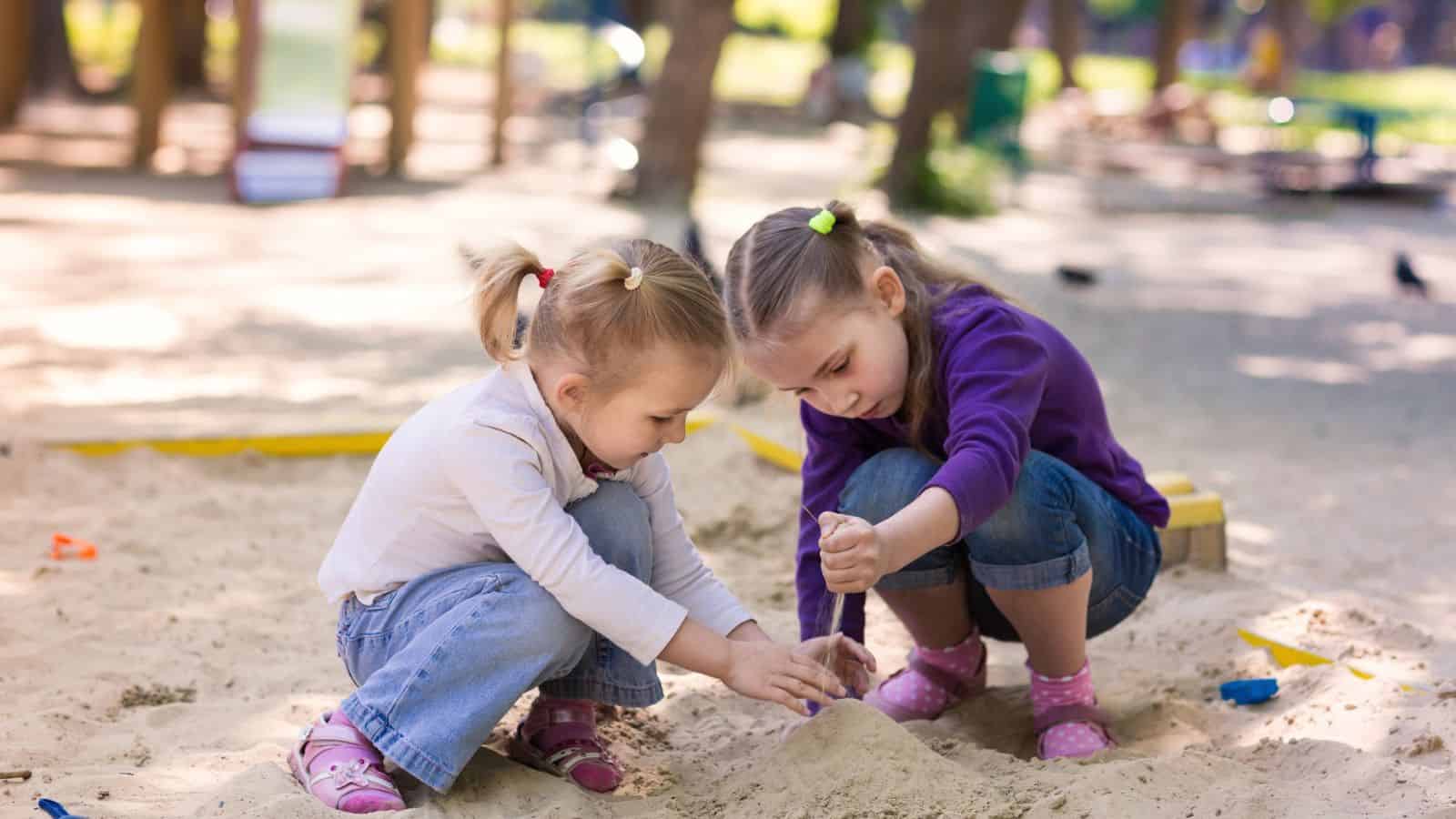 Two young girls playing together and building in a sandbox at a playground on a sunny day.