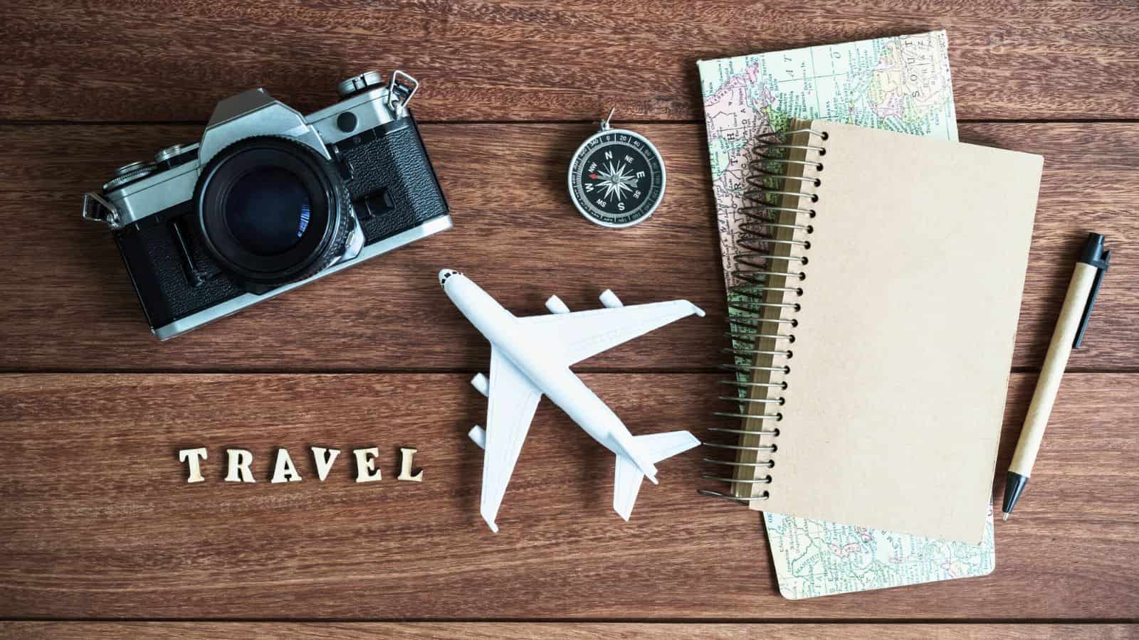 Camera, compass, map, notebook, pen, toy plane, and “TRAVEL” letters arranged on a wooden surface.