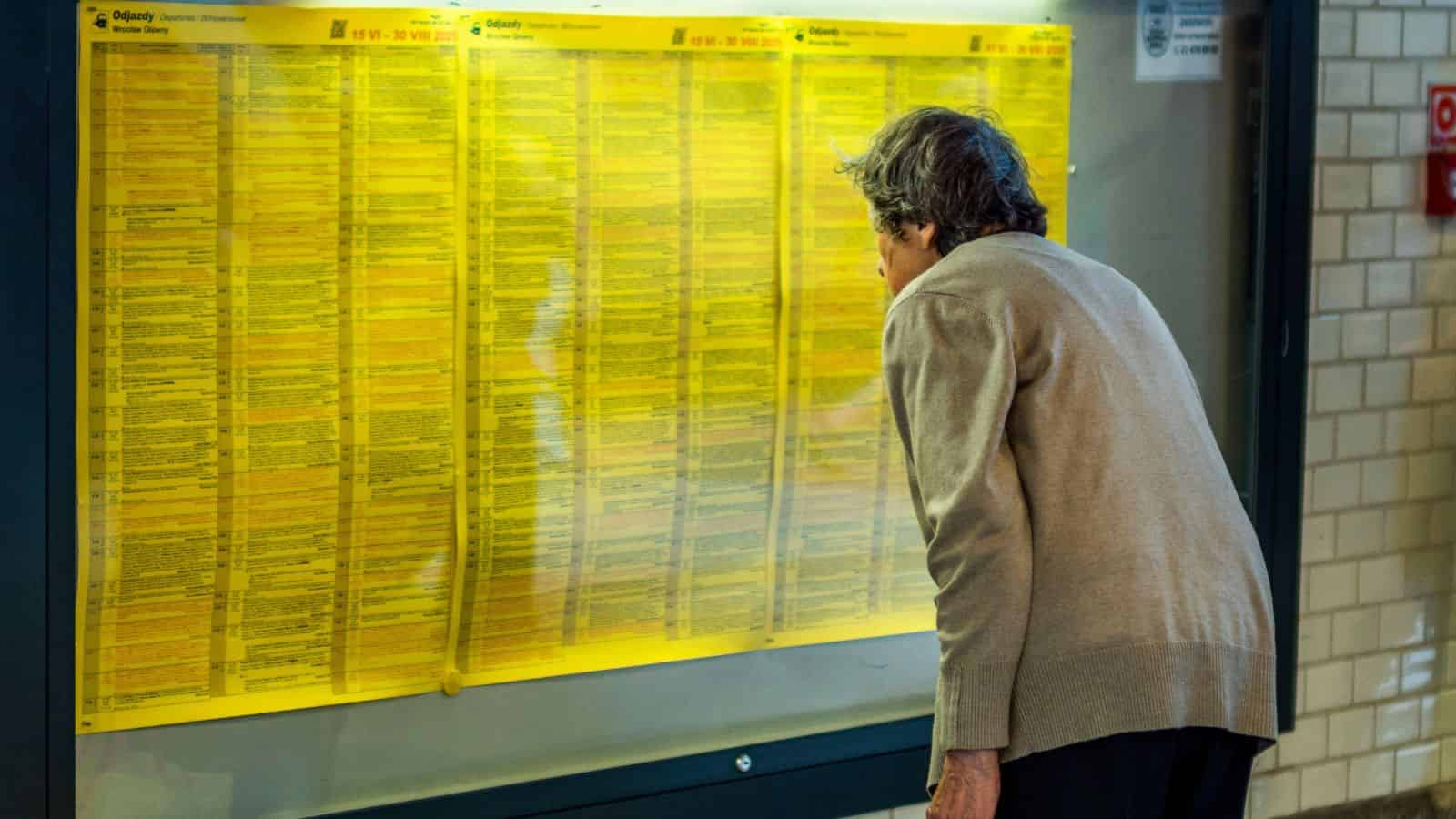 An older woman closely examines a large yellow transit schedule posted on a wall at a station.