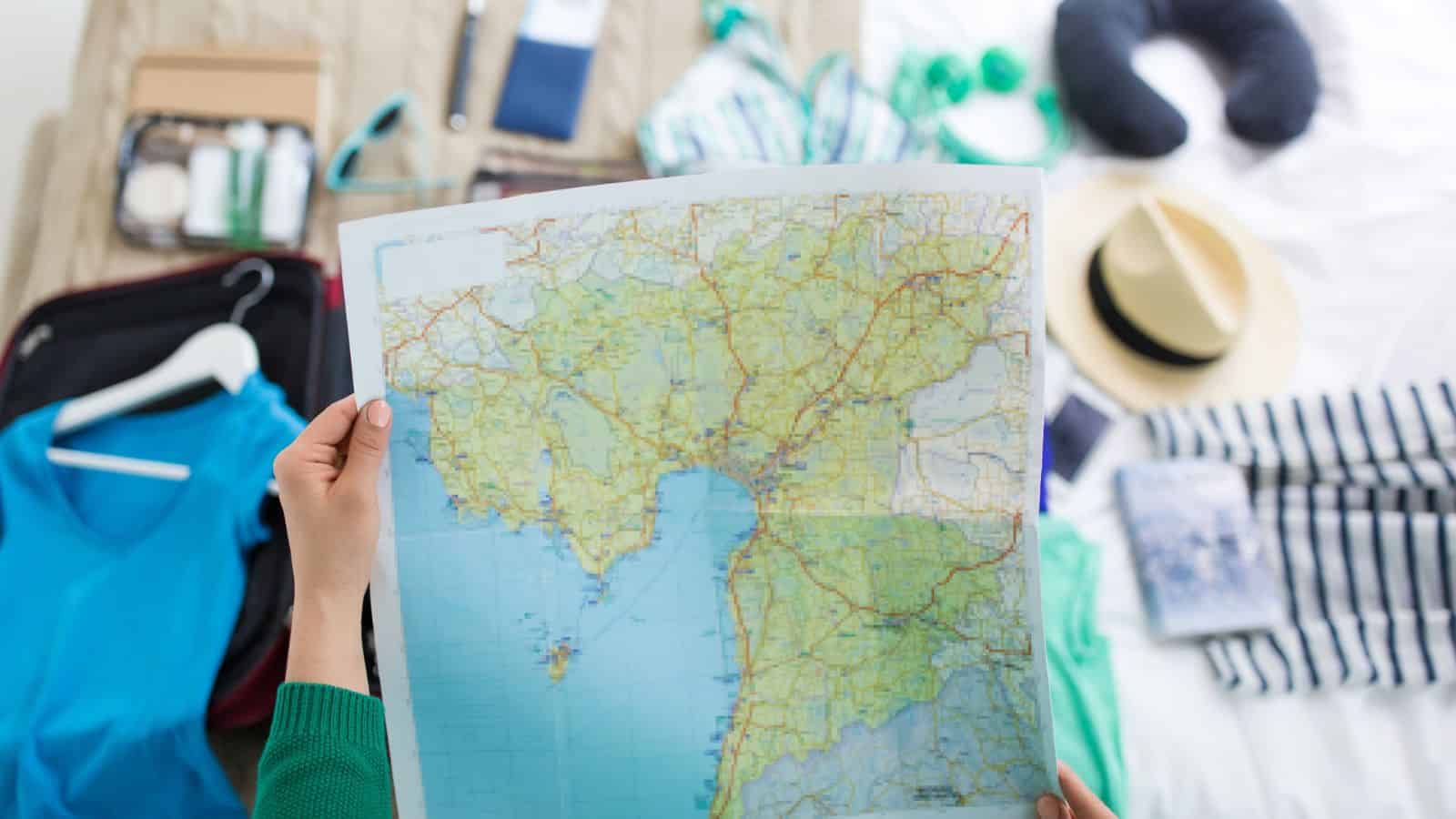 Person holding a map over a bed with travel items, clothes, and accessories spread out.