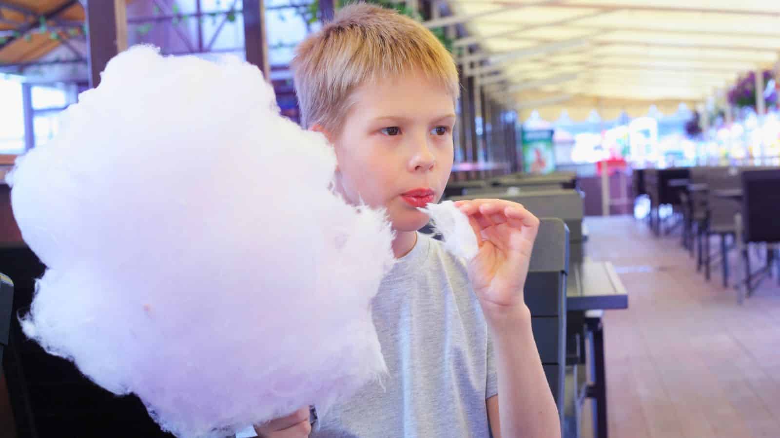 A boy in a gray shirt eats pink cotton candy while sitting at an outdoor table.