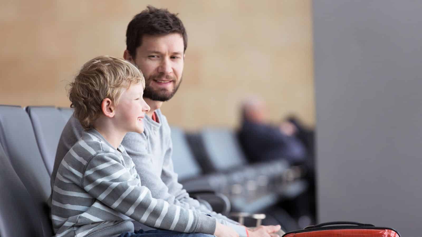 A man and a young boy sit smiling together on chairs in what appears to be an airport waiting area.