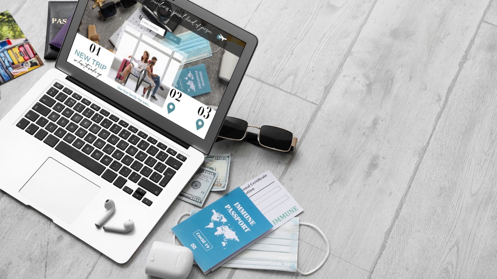 Open laptop with a travel site, passport, money, sunglasses, and mask on a light wooden floor.