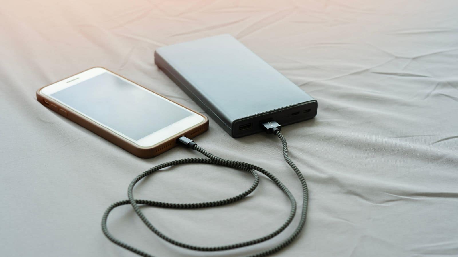 A smartphone connected to a power bank with a braided charging cable on a light gray fabric surface.