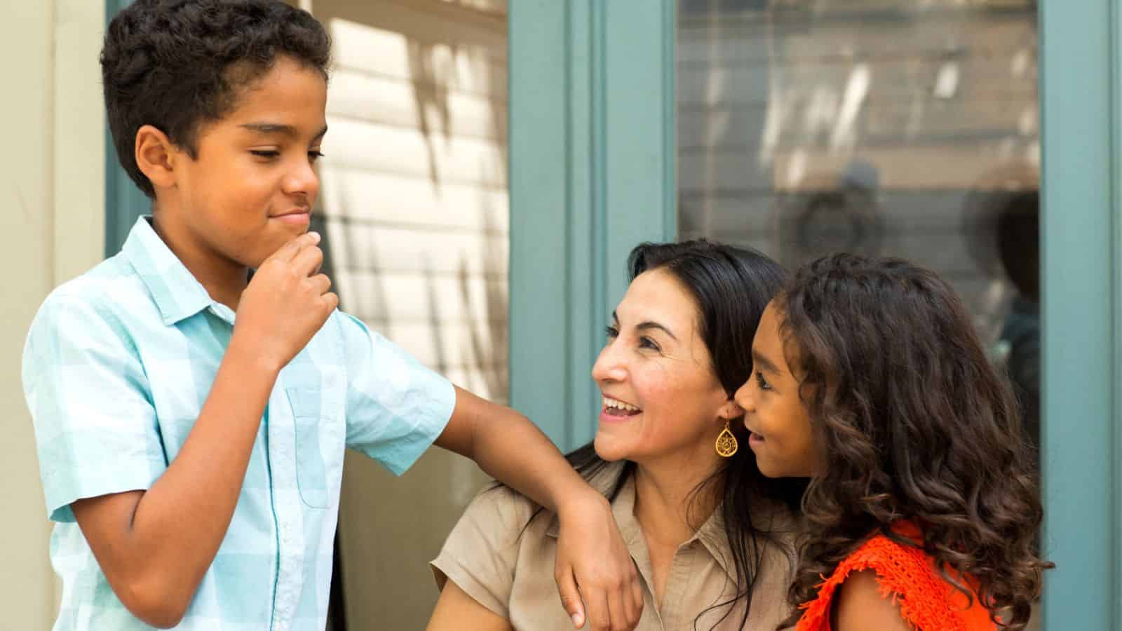 Smiling woman sits between two children, a boy in blue and a girl in orange, all enjoying a happy moment together.