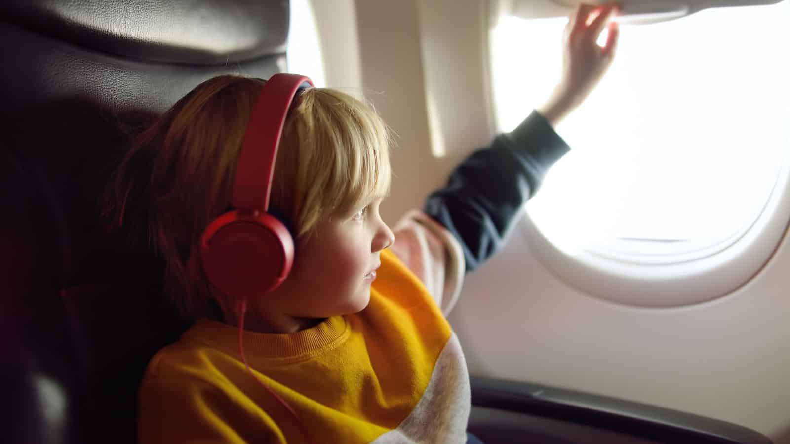 Young child wearing red headphones looks out an airplane window while seated, touching the window shade.