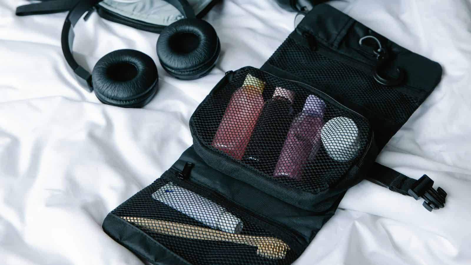 Black travel toiletry bag with bottles, toothbrush, and headphones on a white bed.