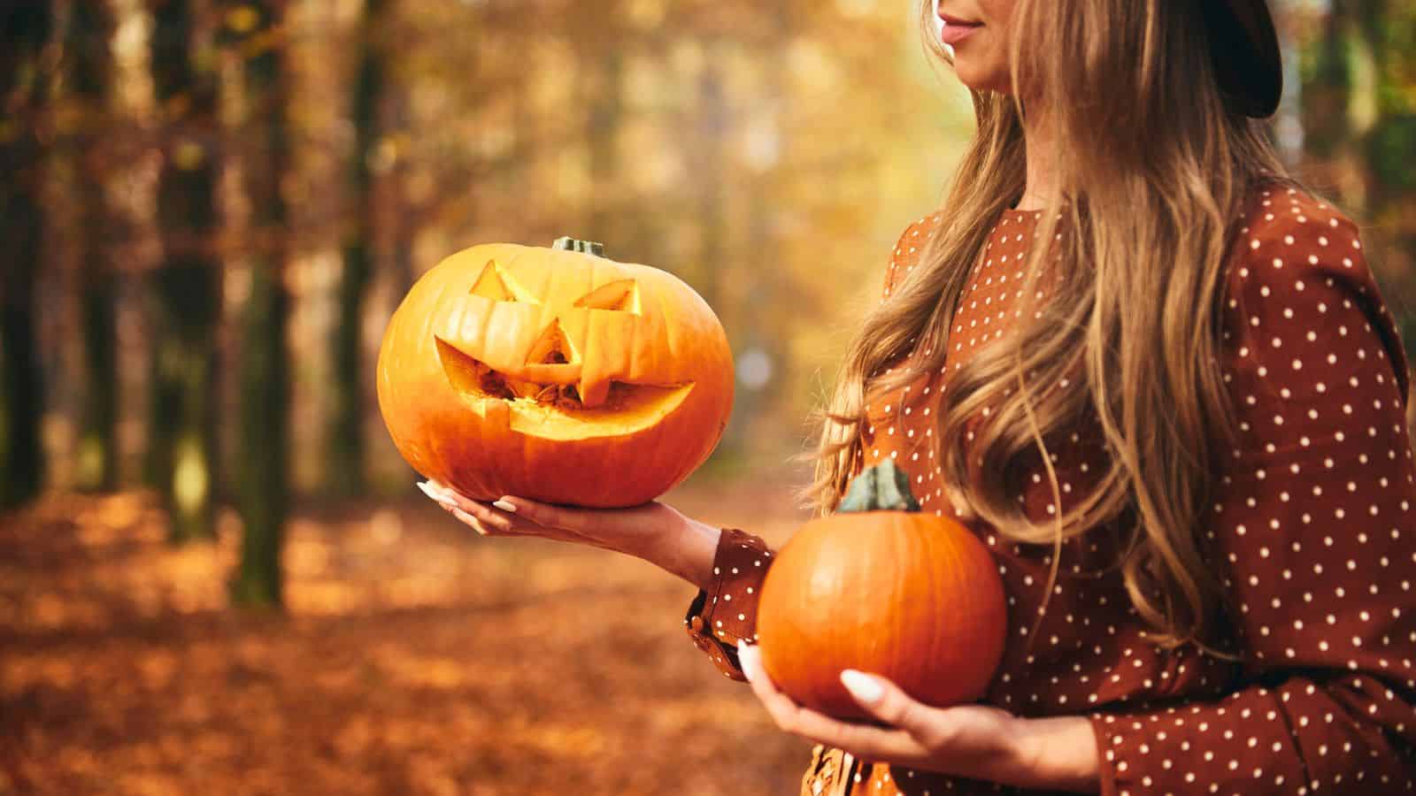 Woman in autumn forest holding a carved jack-o'-lantern pumpkin and an uncarved pumpkin.