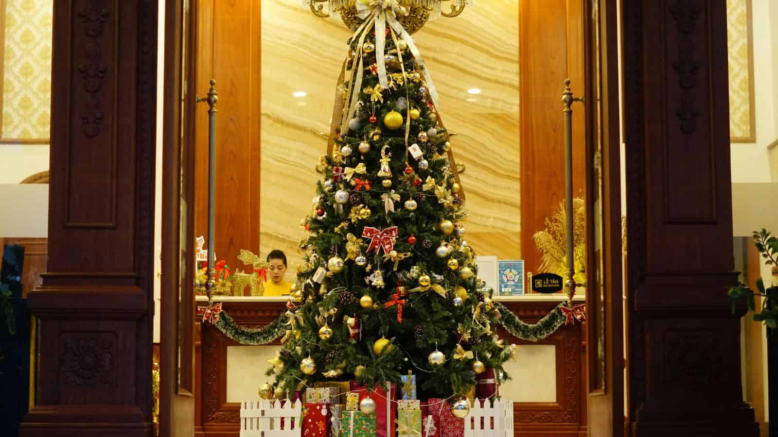 A decorated Christmas tree with ornaments and presents underneath, displayed indoors with a wooden backdrop.