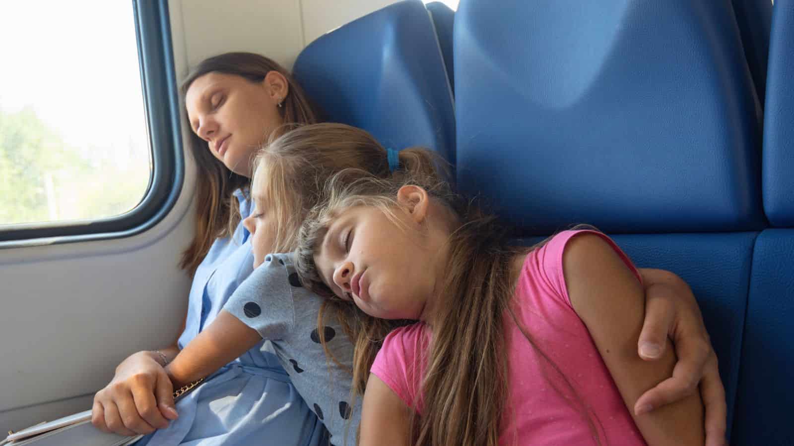 A woman and two girls are sleeping together on blue seats in a train.