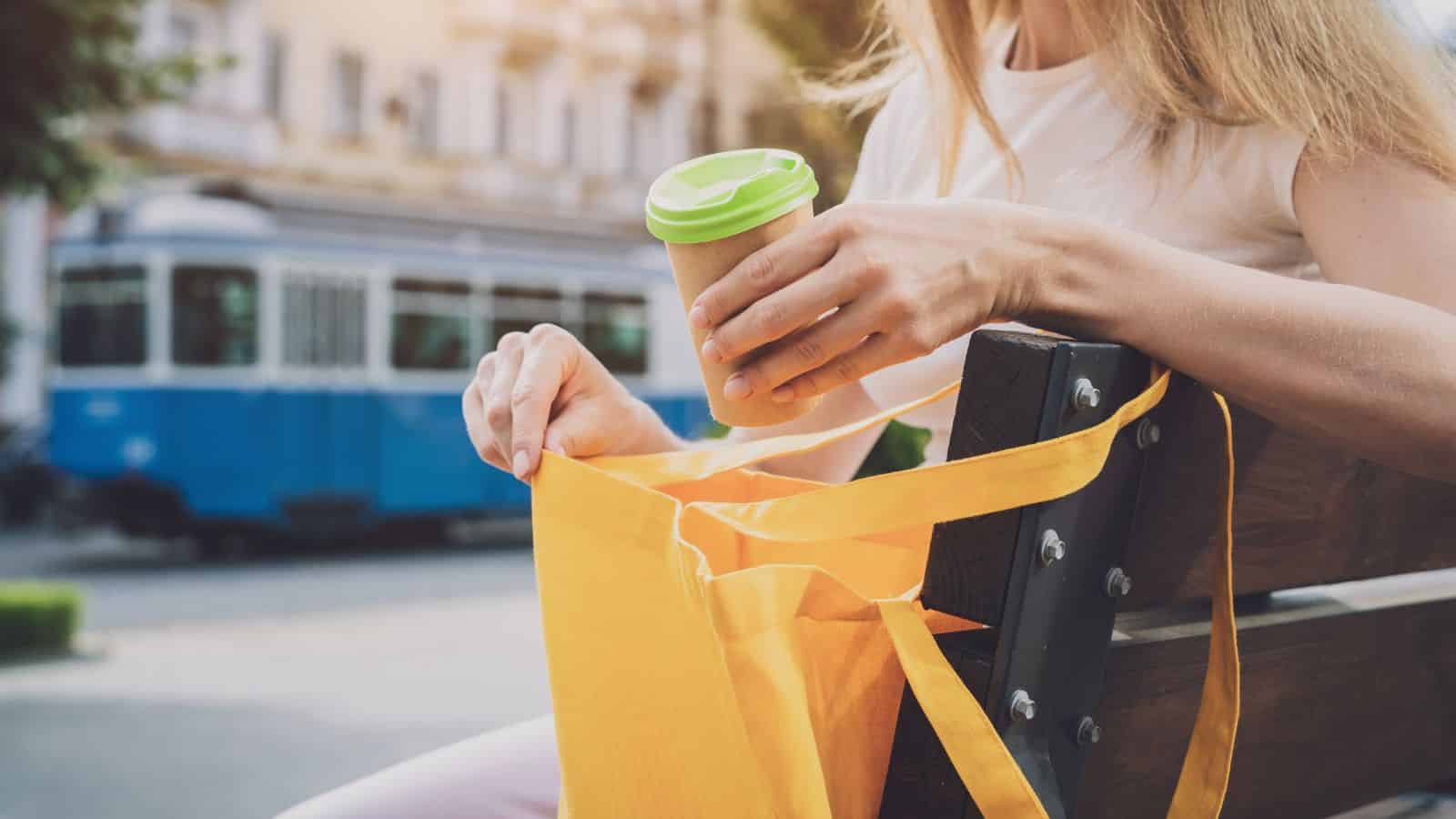 Woman on bench holding a coffee cup and yellow tote bag; blue tram in the background.
