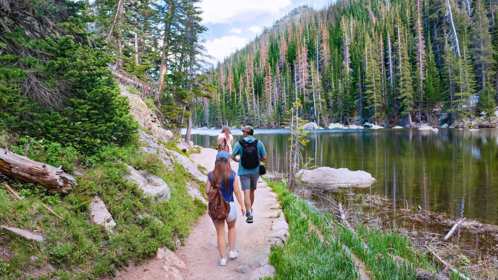 Three people hike on a trail beside a lake surrounded by pine trees and mountains.